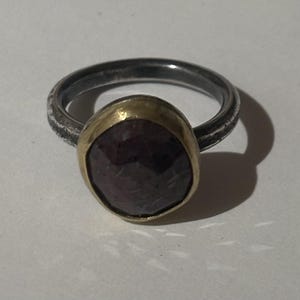May include: A ring featuring a dark, faceted, round gemstone set in a gold-toned bezel. The band is made of a dark, textured, oxidized metal that contrasts with the smooth, polished gold setting surrounding the central stone.