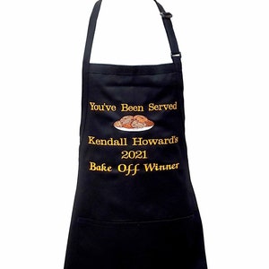 May include: Black apron with a gold embroidered design that says "You've Been Served Kendall Howard's 2021 Bake Off Winner" with a picture of a plate of cookies.