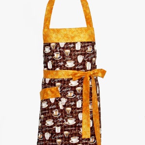 May include: A brown and gold kitchen apron with a coffee cup pattern. The apron has a large pocket on the front and a tie at the waist. The fabric features the words "Coffee", "Java", "Espresso", "Cappuccino", "Latte", and "Mocha".