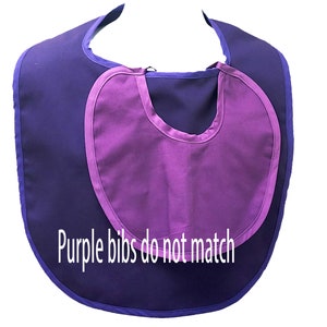 May include: A two-tone purple baby bib with a rounded shape. The bib has a darker purple base with a lighter purple inset and trim. The text "Purple bibs do not match" is printed on the bib.