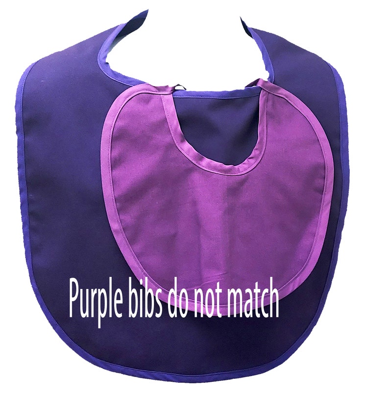 May include: A two-tone purple baby bib with a rounded shape. The bib has a darker purple base with a lighter purple inset and trim. The text "Purple bibs do not match" is printed on the bib.