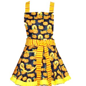 Puede incluir: Un delantal negro y amarillo con estampado de calabaza. El delantal tiene una corbata a rayas amarilla y roja y un volante amarillo alrededor del borde inferior.