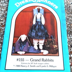 Peut inclure: Une couverture de livre bleue et blanche avec le titre "Dream Spinners" et le num&eacute;ro "#155 - Grand Rabbits". La couverture pr&eacute;sente un dessin de deux lapins blancs v&ecirc;tus de v&ecirc;tements. Le texte sous le titre indique "Patterns for 30" draft stopper rabbits". Les informations de copyright indiquent "1988 Nancy J. Smith and Lynda S. Milligan".
