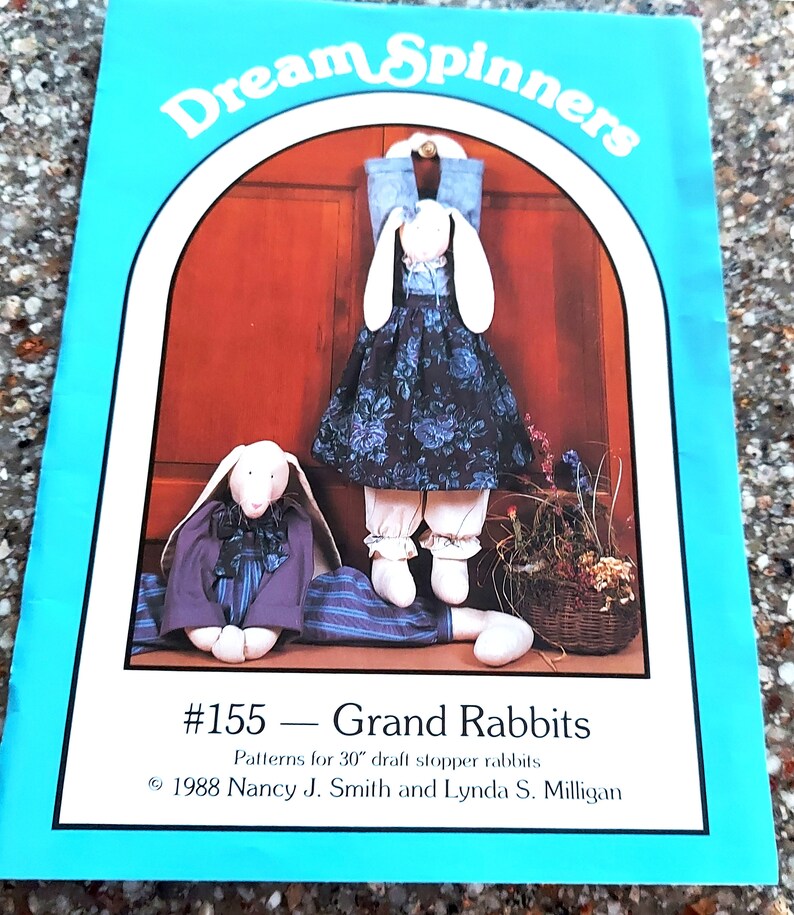Peut inclure: Une couverture de livre bleue et blanche avec le titre "Dream Spinners" et le num&eacute;ro "#155 - Grand Rabbits". La couverture pr&eacute;sente un dessin de deux lapins blancs v&ecirc;tus de v&ecirc;tements. Le texte sous le titre indique "Patterns for 30" draft stopper rabbits". Les informations de copyright indiquent "1988 Nancy J. Smith and Lynda S. Milligan".