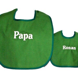 May include: Two green bibs with a dark green trim. The larger bib has the word "Papa" in white, and the smaller bib has the name "Ronan" in white. Both bibs are made of a soft material.