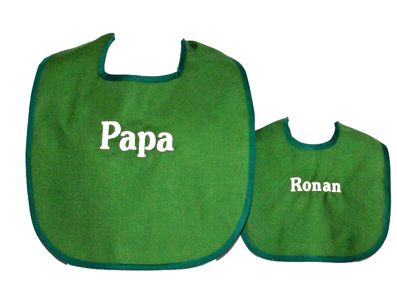May include: Two green bibs with a dark green trim. The larger bib has the word "Papa" in white, and the smaller bib has the name "Ronan" in white. Both bibs are made of a soft material.