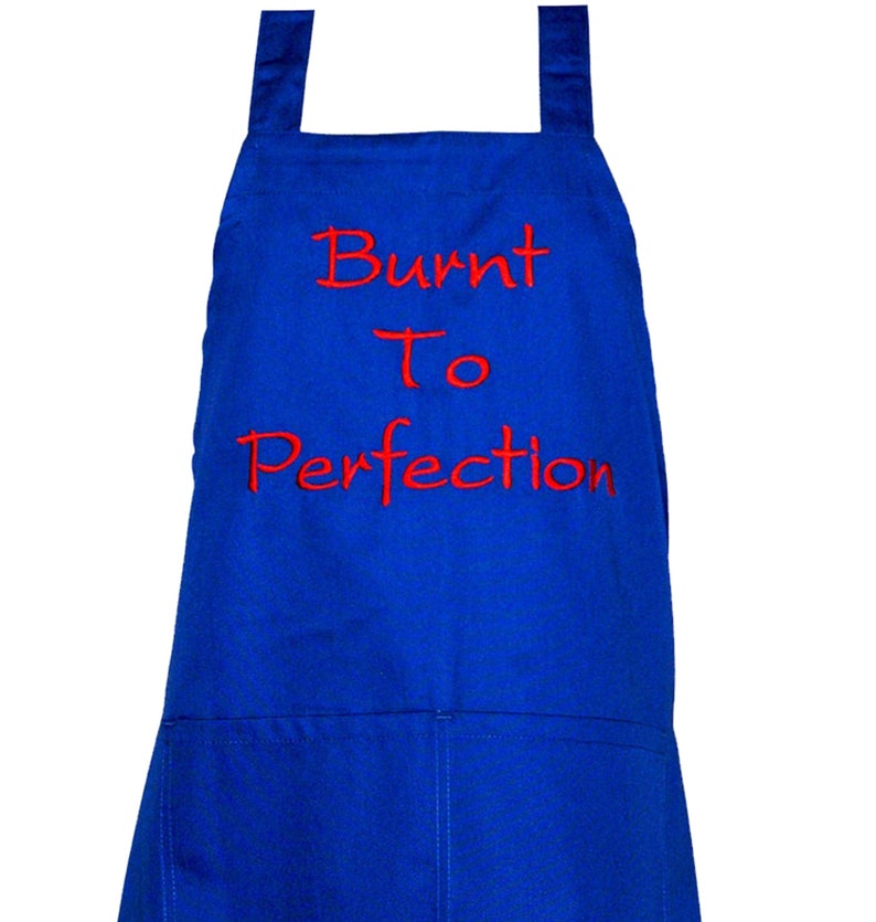 Pode incluir: Um avental azul royal com dois bolsos frontais e al&ccedil;as. O avental tem o texto bordado "Burnt To Perfection" em vermelho. O avental &eacute; projetado para cozinhar e assar.