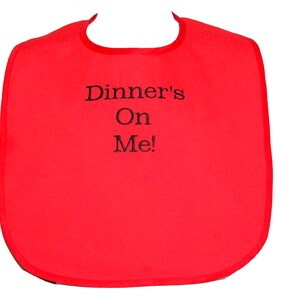 May include: A bright red bib with a rounded bottom and a neck opening. The bib has black embroidered text that reads "Dinner's On Me!" The edges are trimmed with a darker red binding.