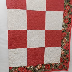 May include: A red and white quilt with a poinsettia print border. The quilt is made of squares with a quilted design of a flower.