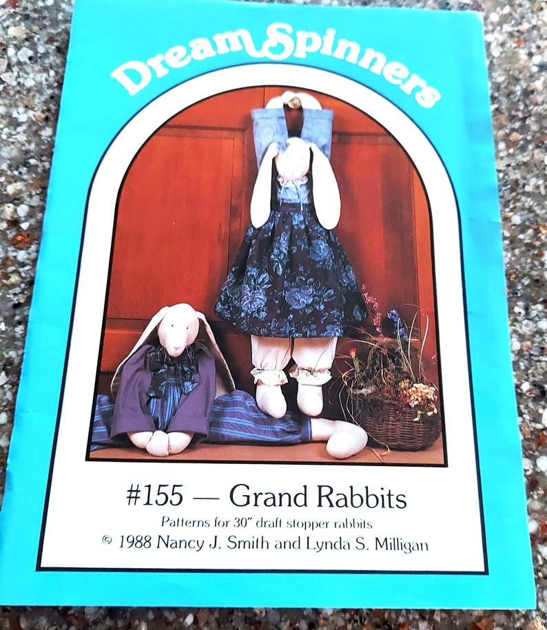 Peut inclure: Une couverture de livre pour un patron de couture intitul&eacute; "Dream Spinners #155 - Grand Rabbits". La couverture pr&eacute;sente un fond bleu avec une arche blanche. Deux poup&eacute;es de lapin blanc sont repr&eacute;sent&eacute;es, l'une portant un gilet en jean bleu et une robe &agrave; fleurs, et l'autre portant une veste et un pantalon violets. Le texte "Patterns for 30" draft stopper rabbits" est imprim&eacute; sous le titre. Les informations de copyright "&copy; 1988 Nancy J. Smith and Lynda S. Milligan" sont imprim&eacute;es sous le texte.