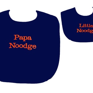 May include: Two navy blue baby bibs. The larger bib has the text "Papa Noodge" in orange, and the smaller bib has the text "Little Noodge" in orange. Both bibs are on a white background.
