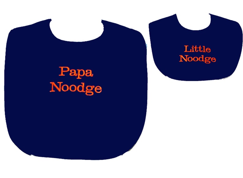 May include: Two navy blue baby bibs. The larger bib has the text "Papa Noodge" in orange, and the smaller bib has the text "Little Noodge" in orange. Both bibs are on a white background.