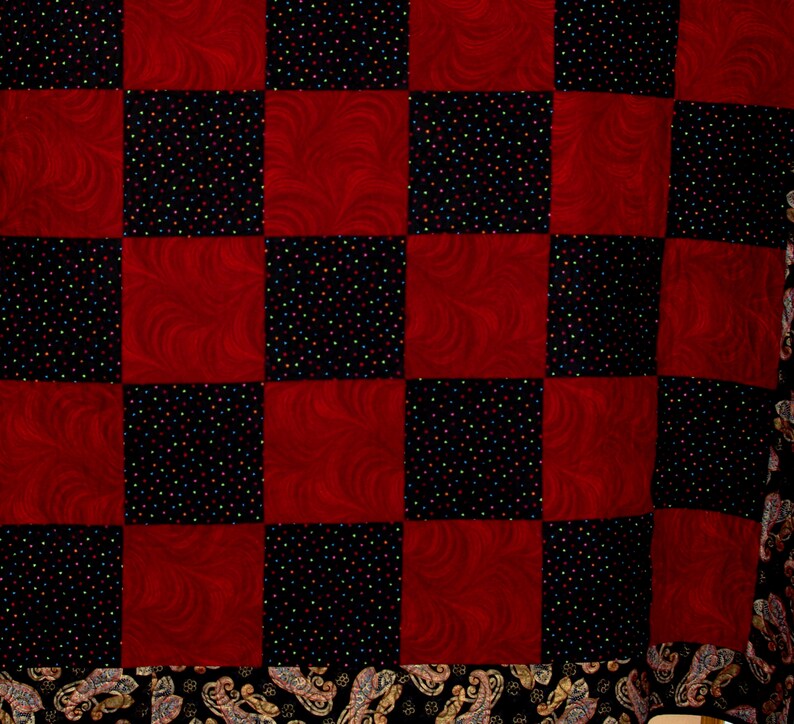 May include: A red and black checkered quilt with a paisley border. The quilt is made of fabric with a swirl pattern and a black fabric with small colorful dots.