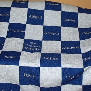 May include: A blue and white checkered quilt with names embroidered in white. The quilt features the words "The Teubert Family" and individual names like "Claudia," "Abigail," and "Lucas." The quilt has a floral patterned border.