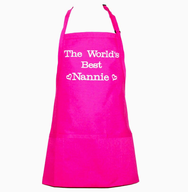 May include: A pink apron with a white pocket and the text "The World's Best Nannie" with a heart on each side of the word "Nannie".