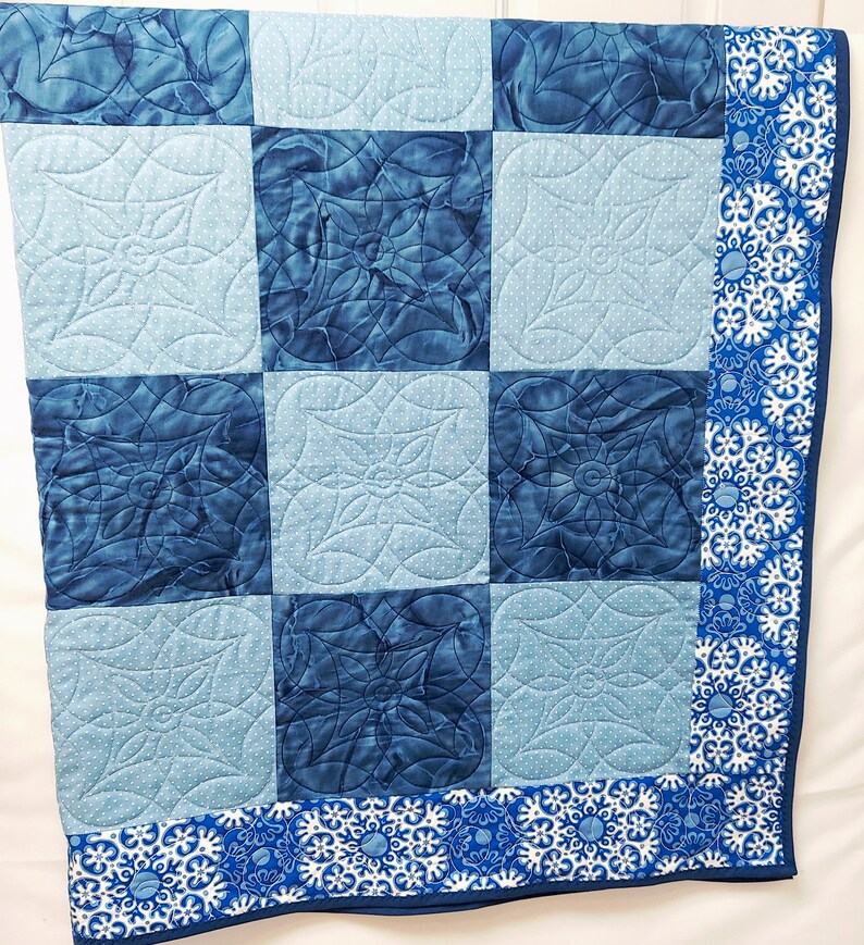 May include: A blue and white quilt with a floral pattern. The quilt is made of squares with different shades of blue and white. The border of the quilt is a white fabric with a blue floral pattern.