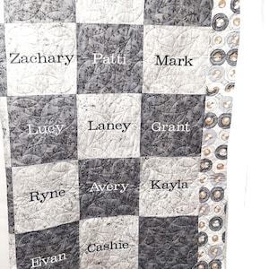 May include: A gray and white quilt with a checkered pattern. Each square has a name embroidered on it, including Abby, Andy, Arie, Zachary, Patti, Mark, Lucy, Laney, Grant, Ryne, Avery, Kayla, Evan, and Cashie.