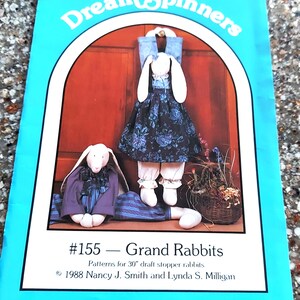 Peut inclure: Une couverture de livre bleue et blanche avec le titre "Dream Spinners" et le num&eacute;ro "#155 - Grand Rabbits". La couverture pr&eacute;sente un dessin de deux lapins blancs v&ecirc;tus de v&ecirc;tements. Le texte sous le titre indique "Patterns for 30" draft stopper rabbits". Les informations de copyright indiquent "1988 Nancy J. Smith and Lynda S. Milligan".