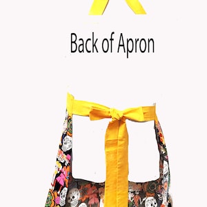 May include: A black apron with a colorful Day of the Dead print featuring skulls, flowers, and other festive imagery. The apron has two yellow ties that tie at the waist.