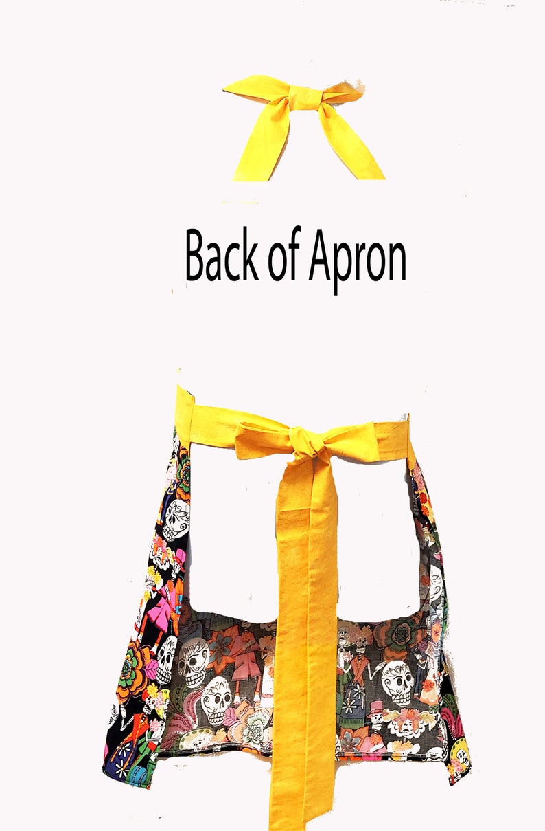 May include: A black apron with a colorful Day of the Dead print featuring skulls, flowers, and other festive imagery. The apron has two yellow ties that tie at the waist.