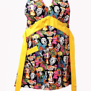 May include: A black halter apron with a yellow ribbon and a colorful Day of the Dead print featuring skeletons, flowers, and skulls.