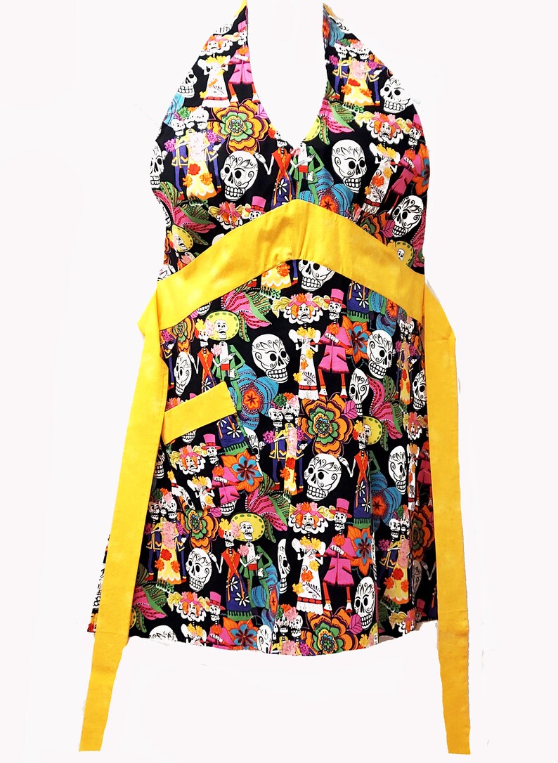 May include: A black halter apron with a yellow ribbon and a colorful Day of the Dead print featuring skeletons, flowers, and skulls.
