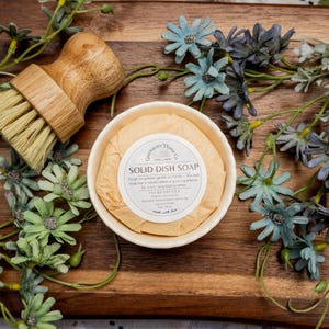May include: A wooden handled dish brush with light bristles and a round solid dish soap in a small bowl. The soap is wrapped in paper with a label that reads "SOLID DISH SOAP". The items are arranged on a wooden cutting board with artificial flowers.