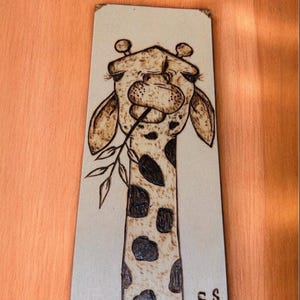 May include: A wood burned image of a giraffe eating a leaf. The giraffe is brown and white with black spots. The image is signed "S.S."