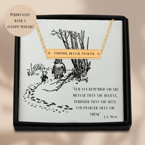 May include: A gold bar necklace with the inscription "Stronger. Braver. Smarter."  The necklace is displayed in a black box with a Winnie the Pooh illustration and a quote from A.A. Milne: "Always remember you are braver than you believe, stronger than you seem, and smarter than you think."
