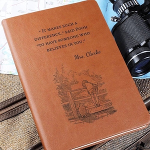 May include: A tan notebook with a smooth cover featuring an engraved illustration of a child and a bear leaning on a wooden fence. The text reads, "It makes such a difference," said Pooh "to have someone who believes in you." and "Mrs. Clarke."
