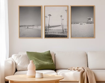 May include: Three black and white framed photographs of a beach scene with palm trees, a pier, and a lifeguard stand. The photos are hanging on a white wall above a white couch.