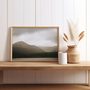 Könnte beinhalten: Ein gerahmtes Foto einer Bergkette mit einem See im Vordergrund. Das Foto hängt an einer weißen Wand über einem Holzregal mit einer weißen Vase und getrockneten Gräsern.