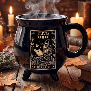 May include: A black ceramic cauldron-shaped mug with three small feet and a large handle. The front features a gold-toned graphic of a person in a witch hat reading a book, with the text OLIVIA THE READER and moon phases above the illustration.