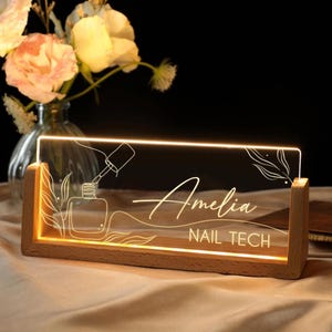 Peut inclure: Panneau lumineux en acrylique avec une base en bois, affichant le nom "Amelia" et "NAIL TECH". Le design comprend une illustration de flacon et de pinceau de vernis à ongles, avec une lumière chaleureuse et accueillante.