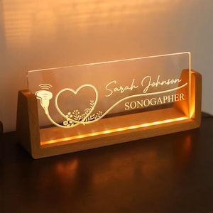 May include: A wooden desk sign with a clear acrylic panel, illuminated from within. The panel displays a heart and ultrasound graphic, the name "Sarah Johnson", and the title "SONOGAPHER". The wooden base emits a warm glow.