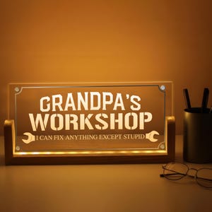 May include: Illuminated sign with the words "GRANDPA'S WORKSHOP" in white, with the phrase "I CAN FIX ANYTHING EXCEPT STUPID" below. The sign is set in a wooden frame and is lit from within, creating a warm glow.