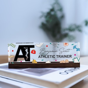 May include: A clear acrylic block with a dark wooden base, featuring the initials "AT" with a medical symbol. The block is personalized with the name "Benjamin Scott" and the title "ATHLETIC TRAINER", along with medical-themed illustrations.