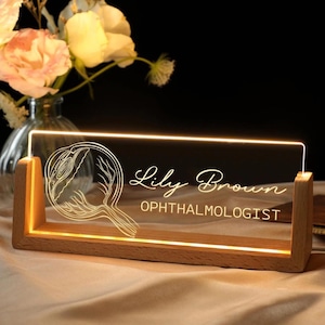 May include: A wooden-based illuminated desk nameplate. The clear acrylic panel features an eye diagram, the name "Lily Brown", and the title "OPHTHALMOLOGIST" in a glowing white font. The rectangular base is made of light-coloured wood.