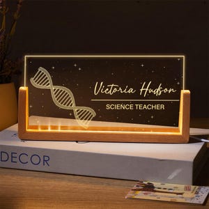 May include: A personalised LED desk lamp with a wooden base. The clear acrylic panel displays a DNA strand graphic, the name "Victoria Hudson", and the title "SCIENCE TEACHER" in a warm, glowing light. The lamp sits on a book.