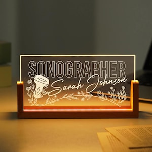 Peut inclure: Un panneau de bureau éclairé avec le mot "SONOGRAPHER" au-dessus du nom "Sarah Johnson". Le panneau a une base en bois et un panneau en acrylique transparent avec un motif floral et un graphique de sonde d'échographie. Le panneau est éclairé d'une lumière chaude.