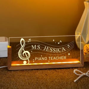 May include: A rectangular, illuminated desk sign with a wooden base. The clear acrylic panel displays a treble clef, musical notes, and the text "MS. JESSICA PIANO TEACHER" in white. The sign emits a warm, yellow glow.