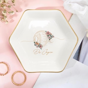 May include: A white, hexagonal trinket dish with a gold rim. The dish features a floral eye design and the text "Dr. Joyce" in gold script. Two gold rings are visible in the lower left corner.