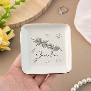 May include: A square, white ceramic trinket dish with a gold rim. The dish features a DNA strand design with floral accents and the name "Camelia" in script. Surrounding the DNA design are several molecular structures.