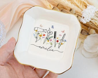 May include: A white, square-shaped trinket dish with a gold rim. The dish features a floral design with a hairdryer, scissors, comb, and a knife. The name "amelia" is written in cursive script.