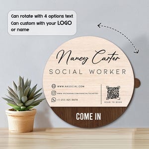 May include: A round wooden sign with the name "Nancy Carter" and the title "Social Worker" in black script. The sign includes contact information, a QR code, and the words "Come In". The sign is light wood with a dark wood bottom.