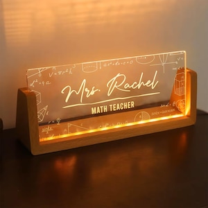 Peut inclure: Un présentoir de bureau éclairé avec une base en bois et une façade en acrylique transparent. L'acrylique affiche le nom "Mrs. Rachel" et "MATH TEACHER" en écriture cursive, ainsi que des formules mathématiques. Un cadeau pour un professeur.