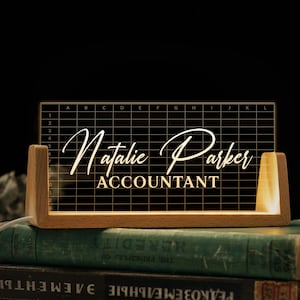 May include: A desk nameplate with a grid design, featuring the name "Natalie Parker" and the title "ACCOUNTANT" in a decorative font. The illuminated nameplate is set in a wooden base.