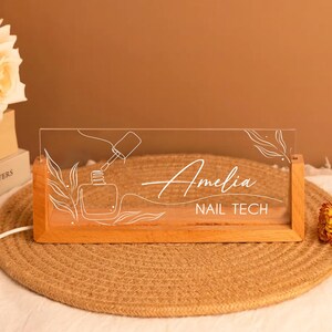 May include: A wooden-based LED sign with the name "Amelia" and the text "NAIL TECH" in white script. The clear acrylic panel features a nail polish bottle and brush design. The sign is illuminated and sits on a woven mat.