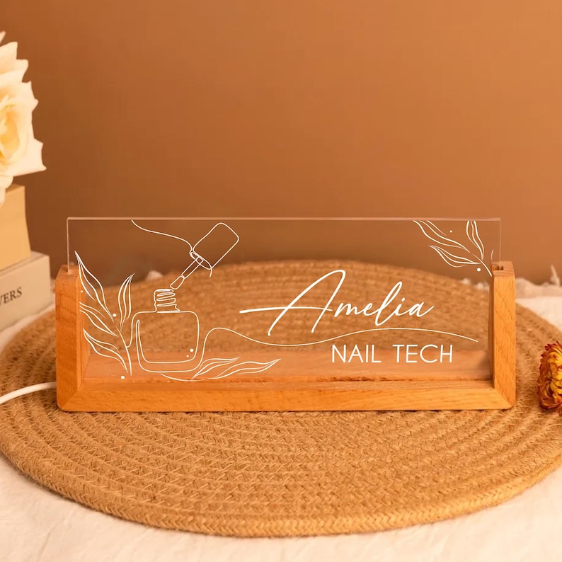 May include: A wooden-based LED sign with the name "Amelia" and the text "NAIL TECH" in white script. The clear acrylic panel features a nail polish bottle and brush design. The sign is illuminated and sits on a woven mat.