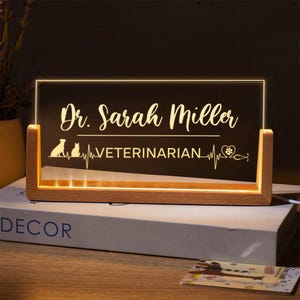 May include: A wooden illuminated sign displaying "Dr. Sarah Miller" in a stylish font. The design includes a dog and cat silhouette, a heartbeat line, and the word "VETERINARIAN". The sign emits a warm, inviting light.